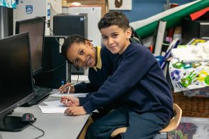 Two young boys in primary school uniforms smiling in front of computers in homework club